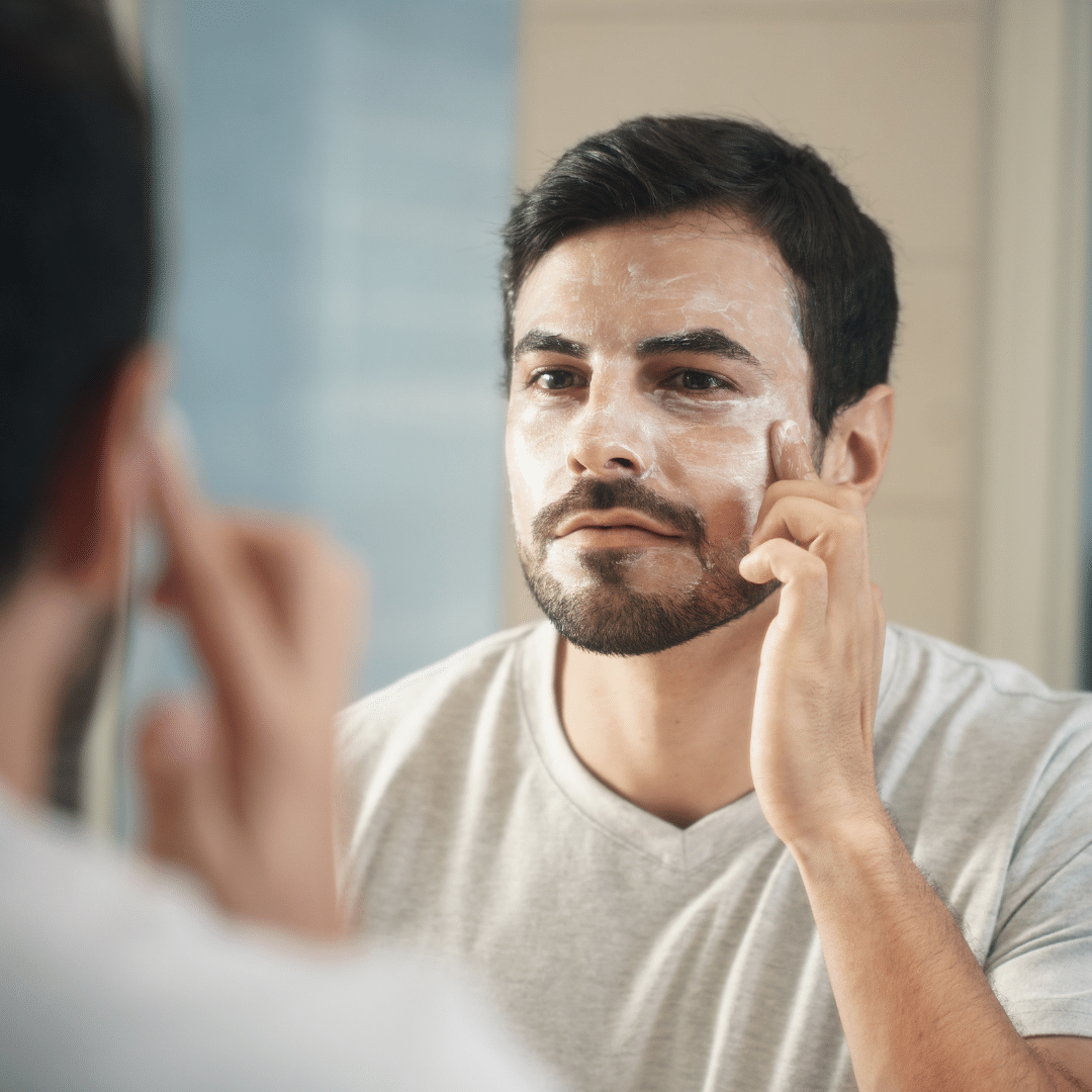 Hombre arreglándose frente al espejo. El uso diario de protector solar y crema hidratante de calidad mantiene tus fibroblastos y colágeno en forma. Ambos productos preservan la barrera protectora de las capas cutáneas superficiales. Y previenen la expansión de procesos inflamatorios inadvertidos a capas profundas de la piel.