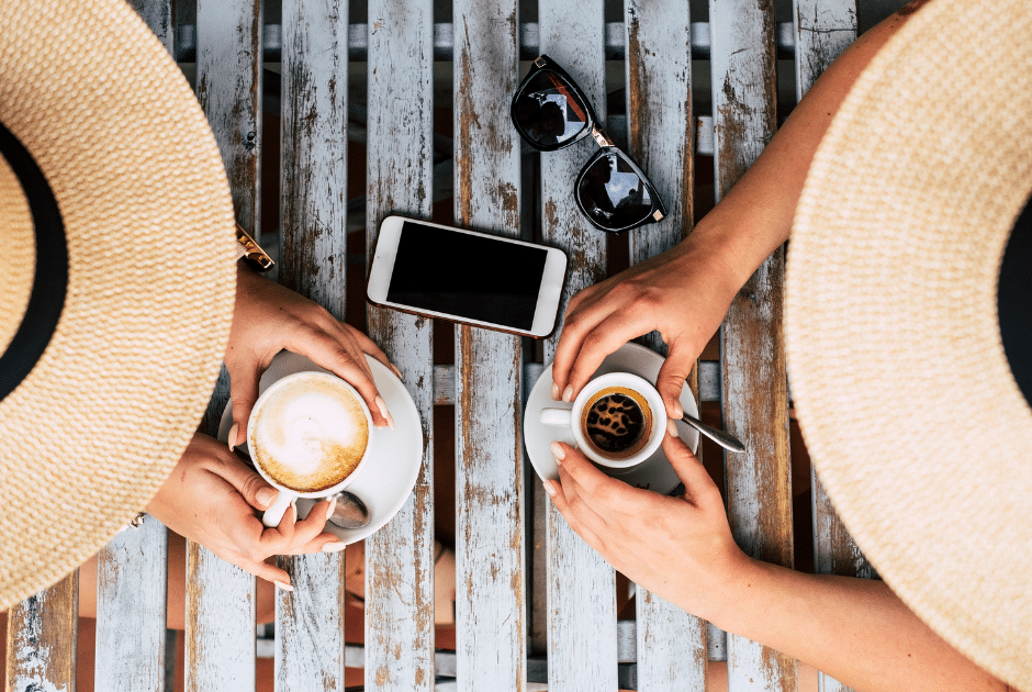 Fotografía de dos personas tomando café al aire libre. Además llevan sombreros para protegerse de la luz solar.
